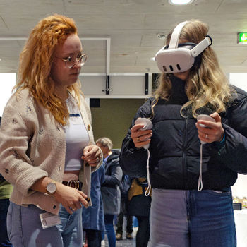 Zwei Frauen stehen nebeneinander, eine trägt eine VR-Brille und hält entsprechende Controller in der Hand.