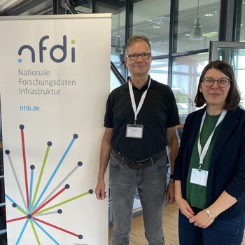Zwei Frauen und ein Mann stehen neben einem Roll-up-Banner, auf welchem das Logo des NFDI abgebildet ist.